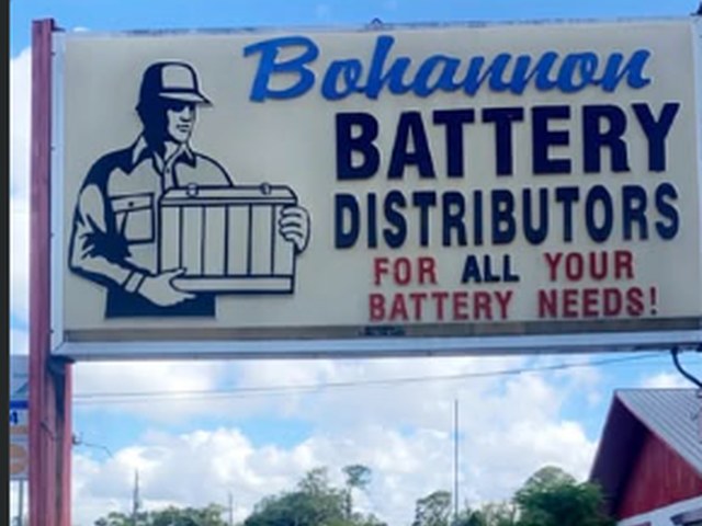The Bohannon Battery Distributors roadside sign on N Palm Ave: a uniformed man carrying a battery beside the words "For all your battery needs!"
